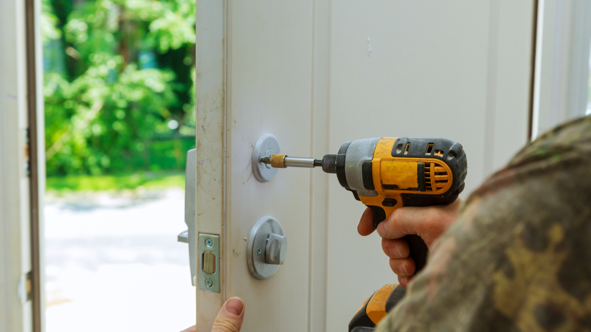 Person using a drill to install a door lock on a white door, outdoors.
