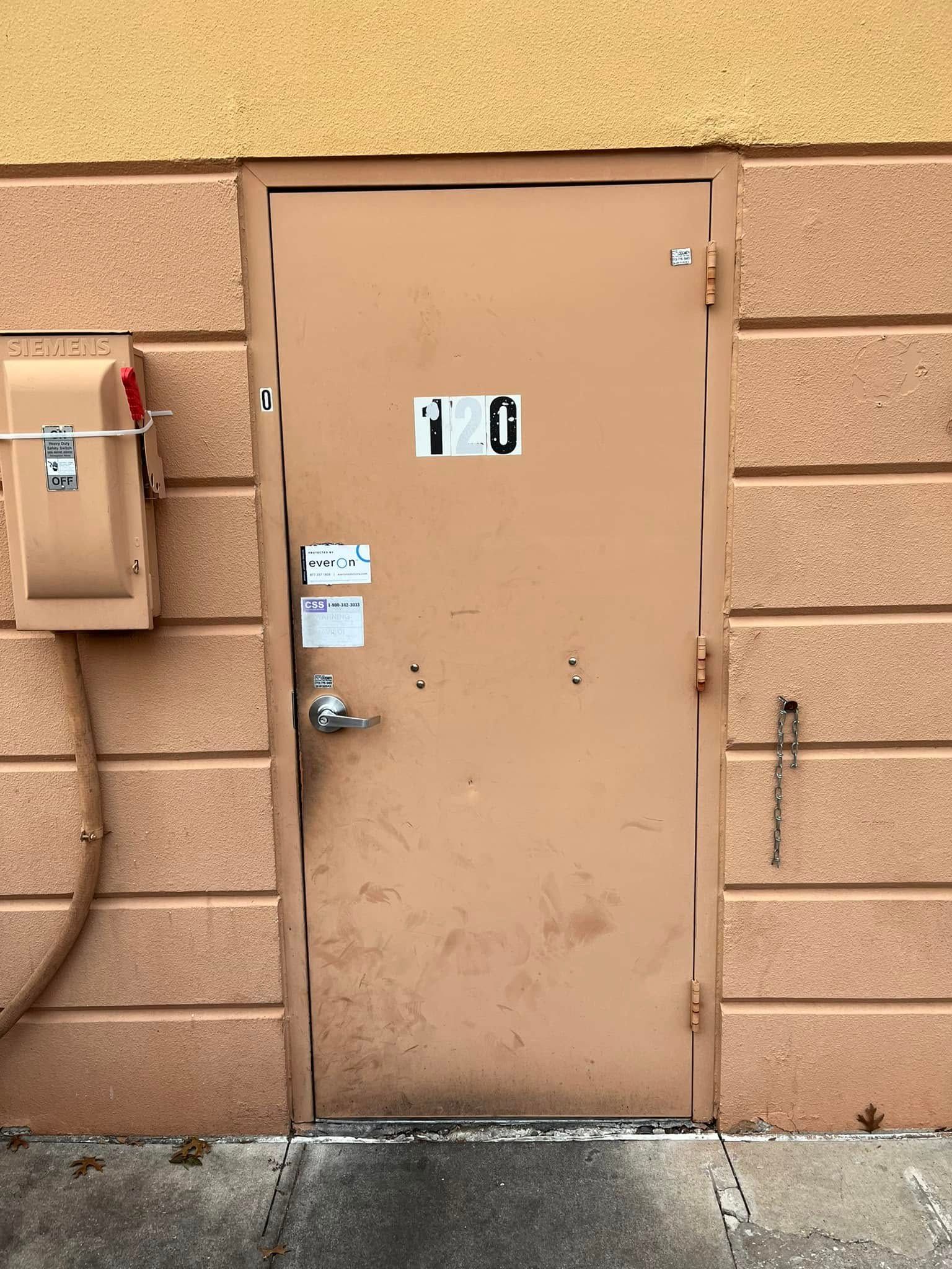 Tan metal door in a brick wall, with a small sign and a box on the side.