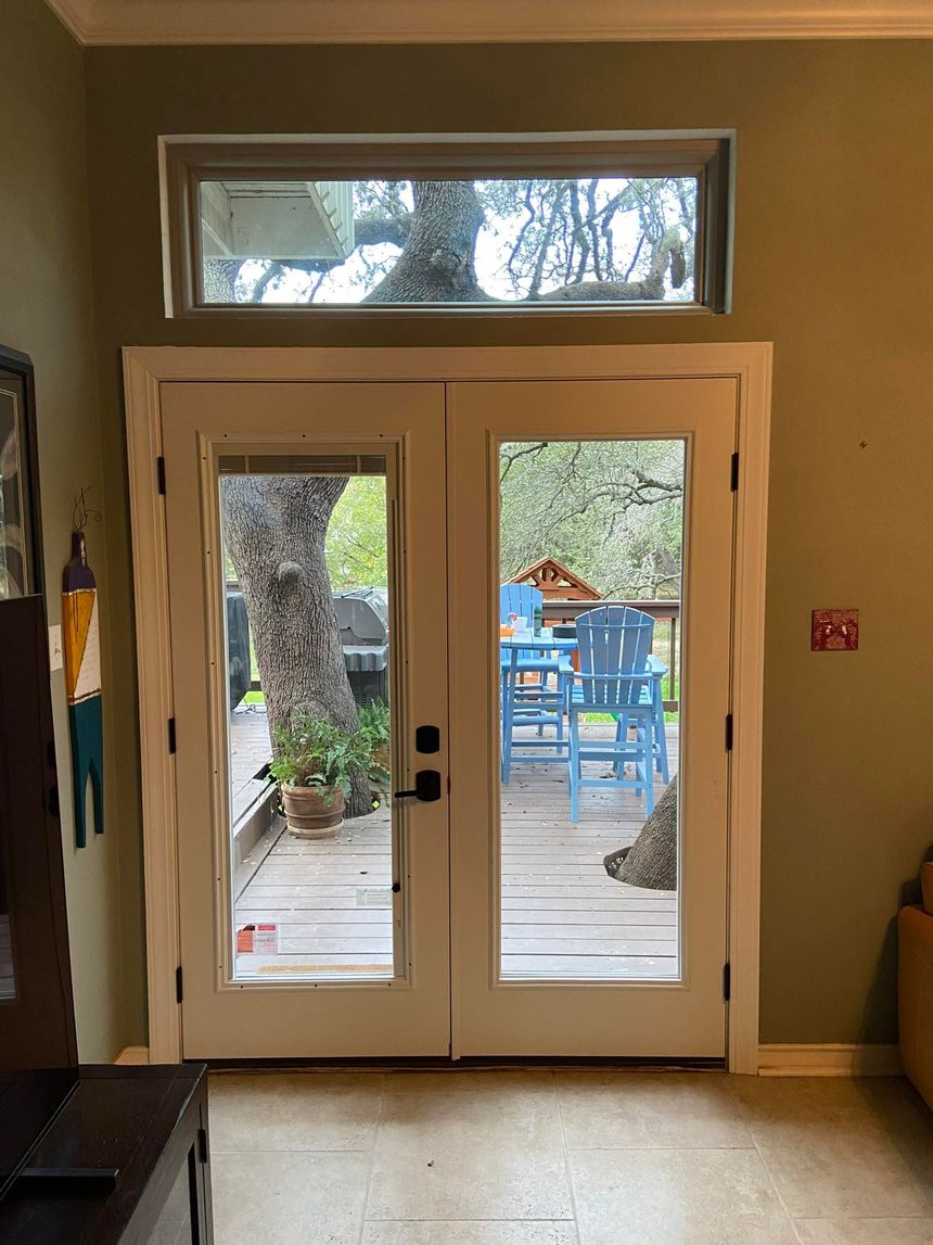 White double doors with glass panels and a small rectangular window above, leading to a deck with blue chairs and a tree.