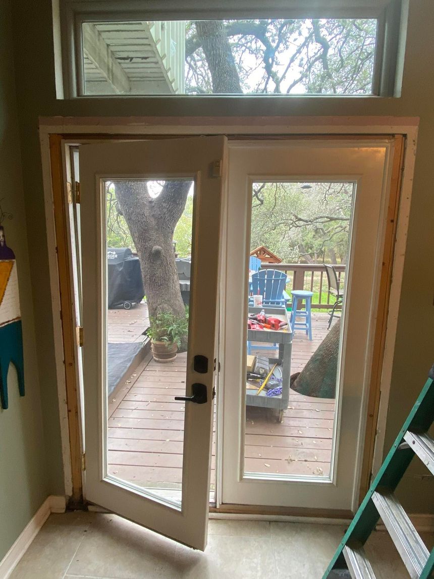 French doors open to a deck with a tree. Door frame and trim are being installed. Green ladder at right.