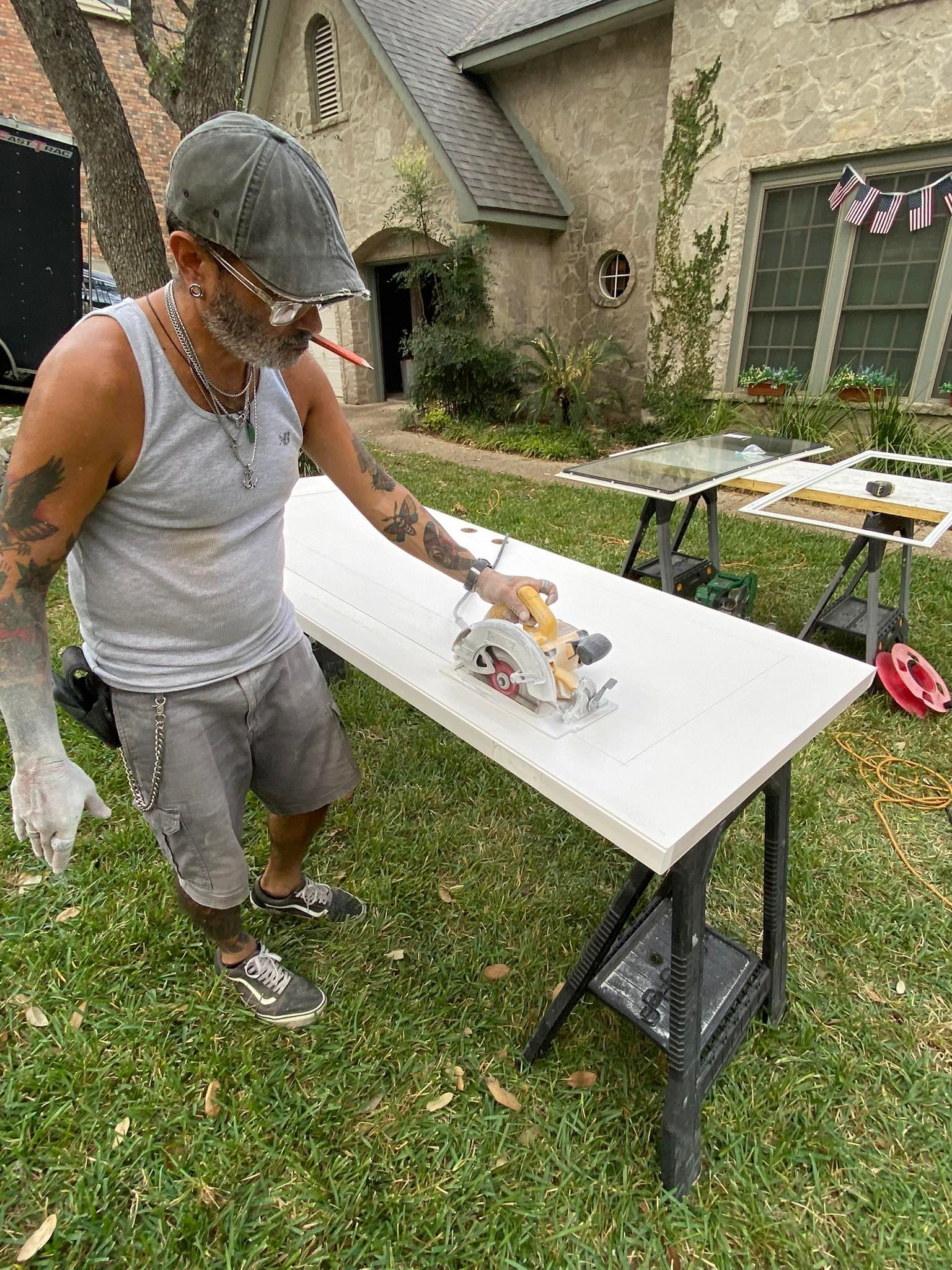 Man using a circular saw on a white countertop outdoors, near a stone house.