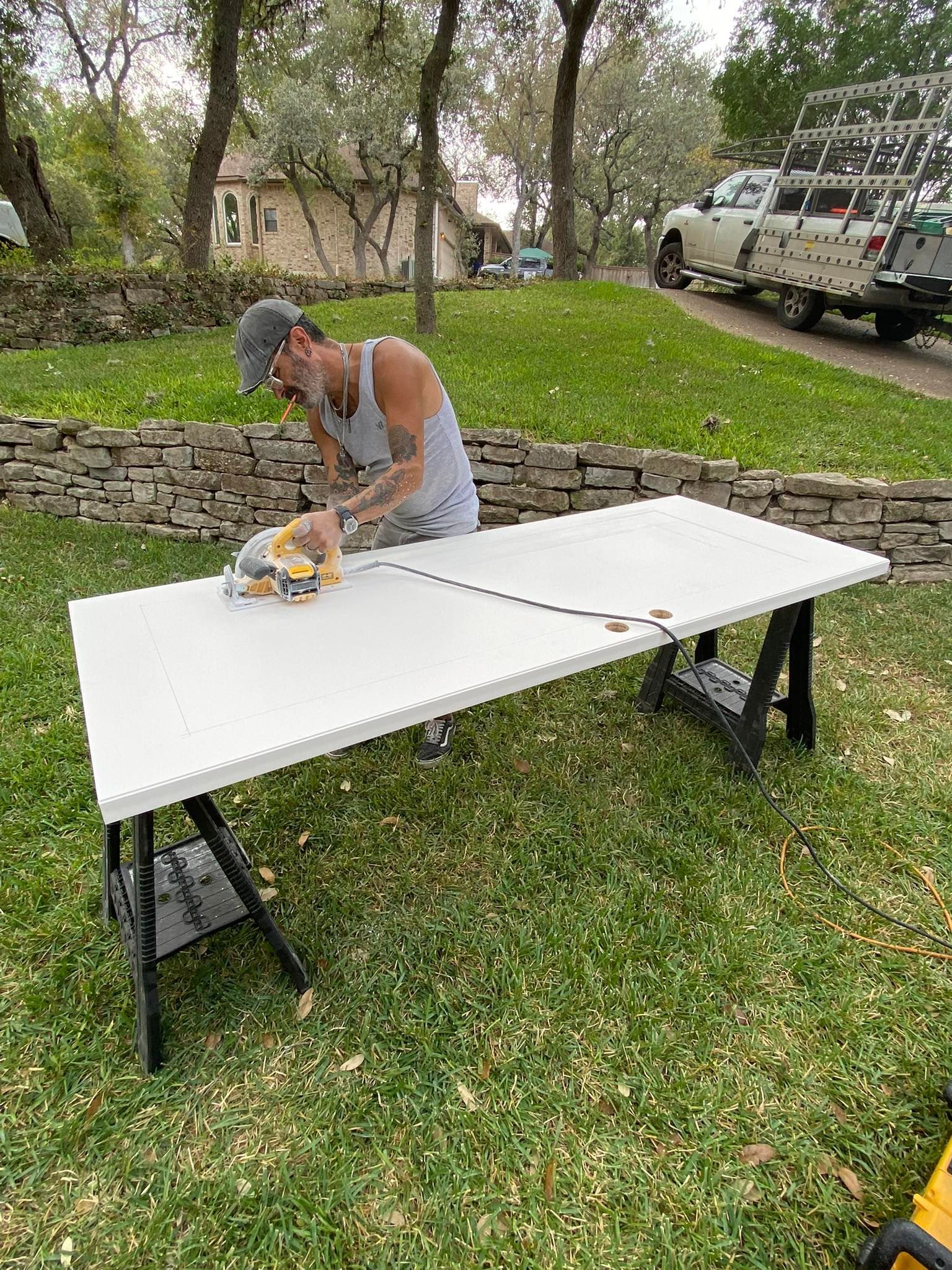 Person uses a saw on a white table set up in a grassy yard. A truck and stone wall are in the background.