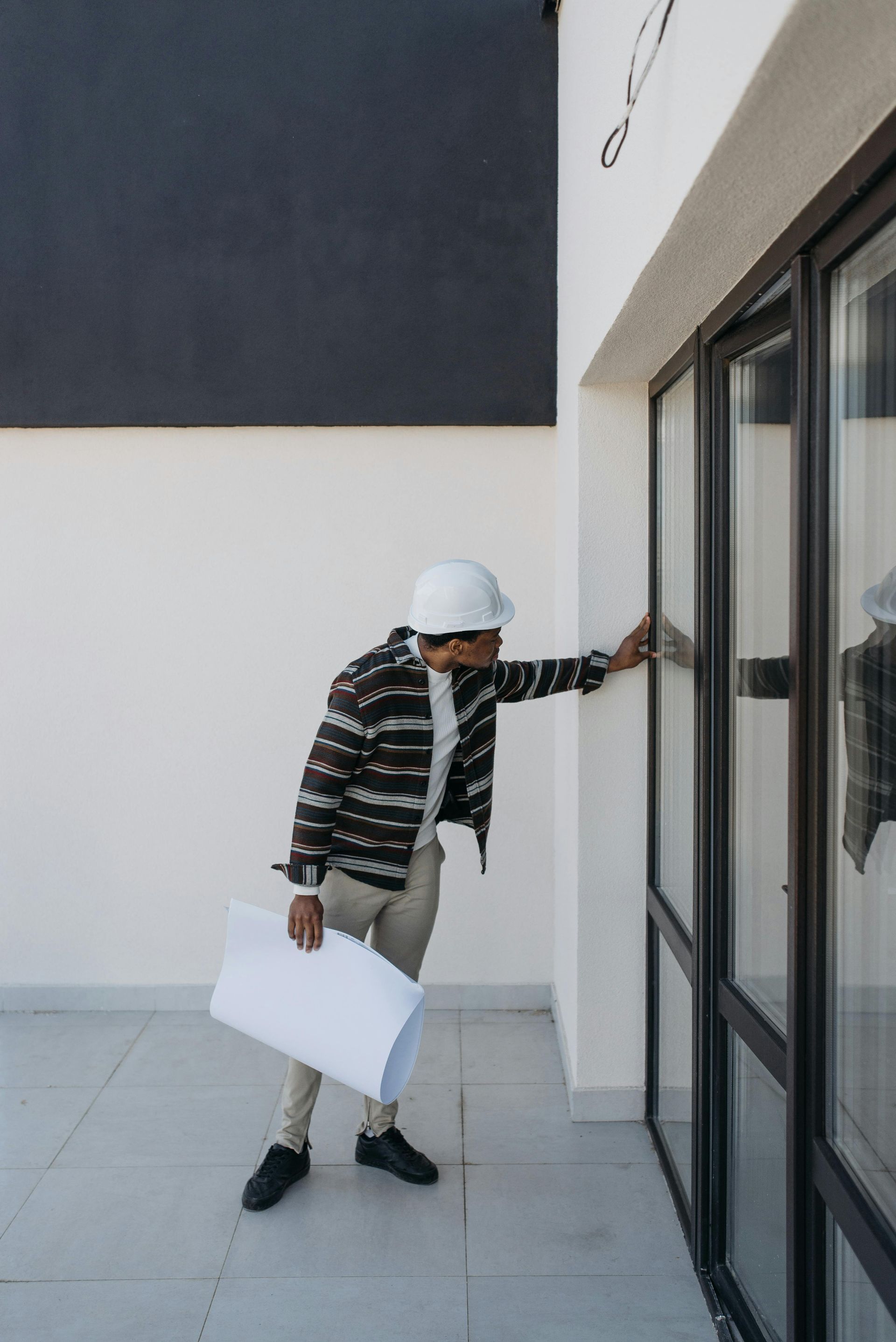 Person in a hard hat inspecting a glass door on a balcony, holding rolled plans.