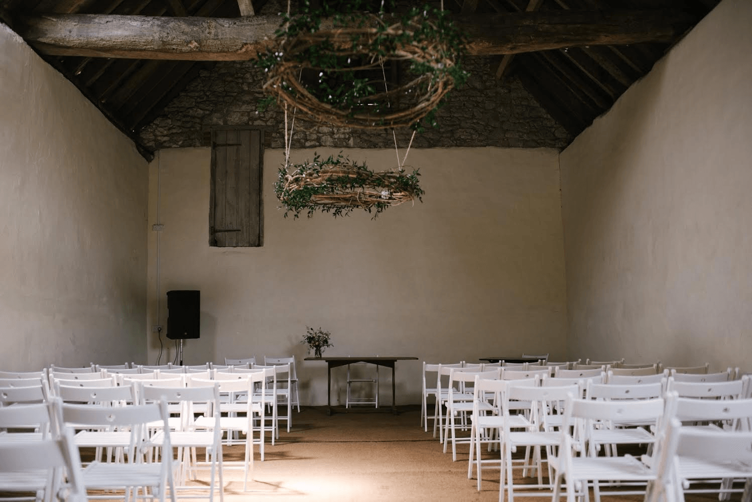 White chairs line a room with floral wreaths hanging from the wooden ceiling, set for an event.