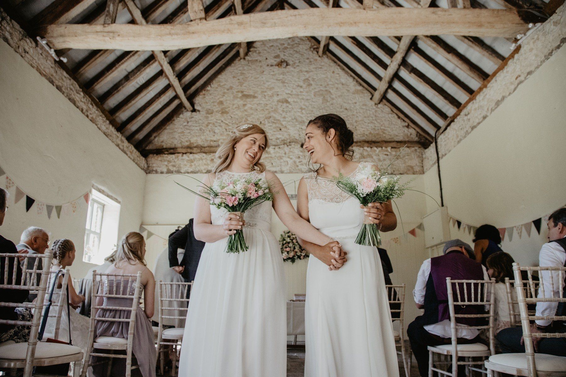 Two brides in white dresses hold hands, smiling, in a barn-like wedding venue with guests.