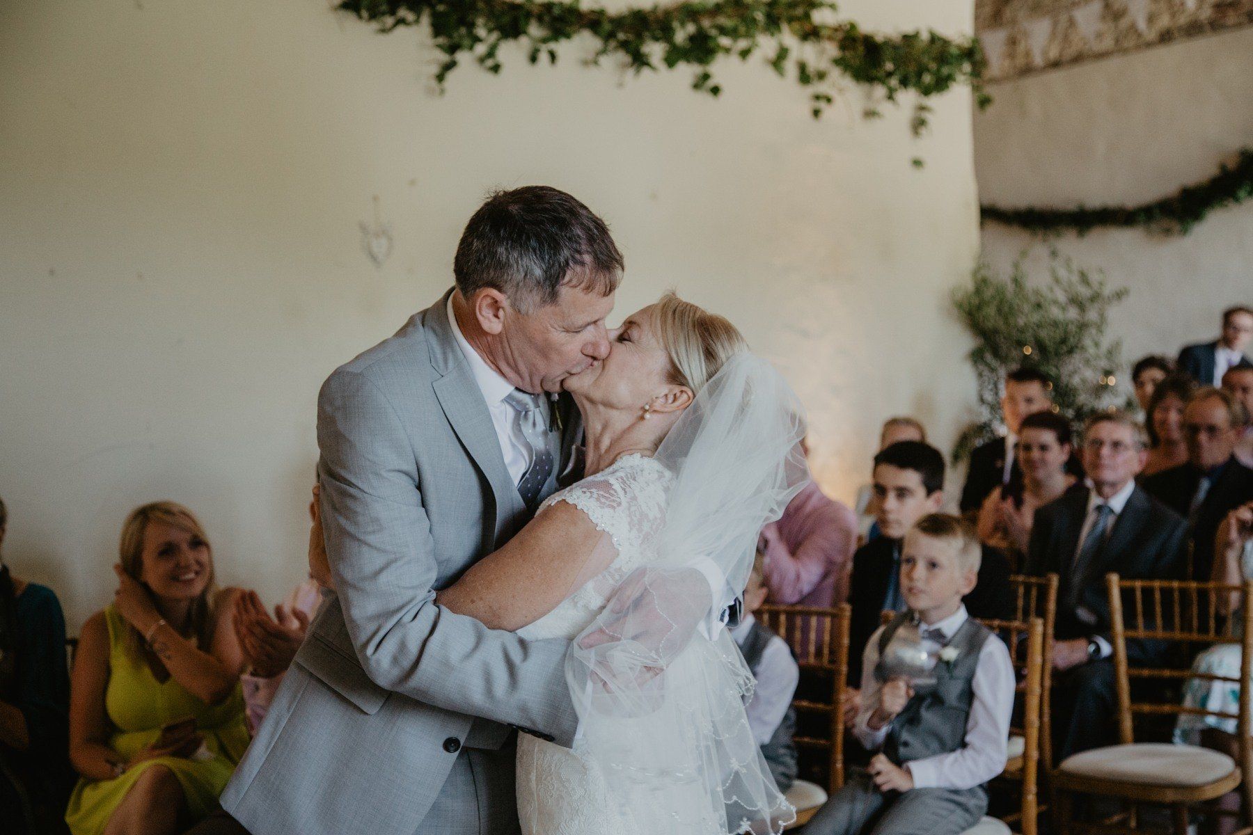 Couple kissing at a wedding ceremony; guests watch. Stone wall backdrop, greenery, sunlight.