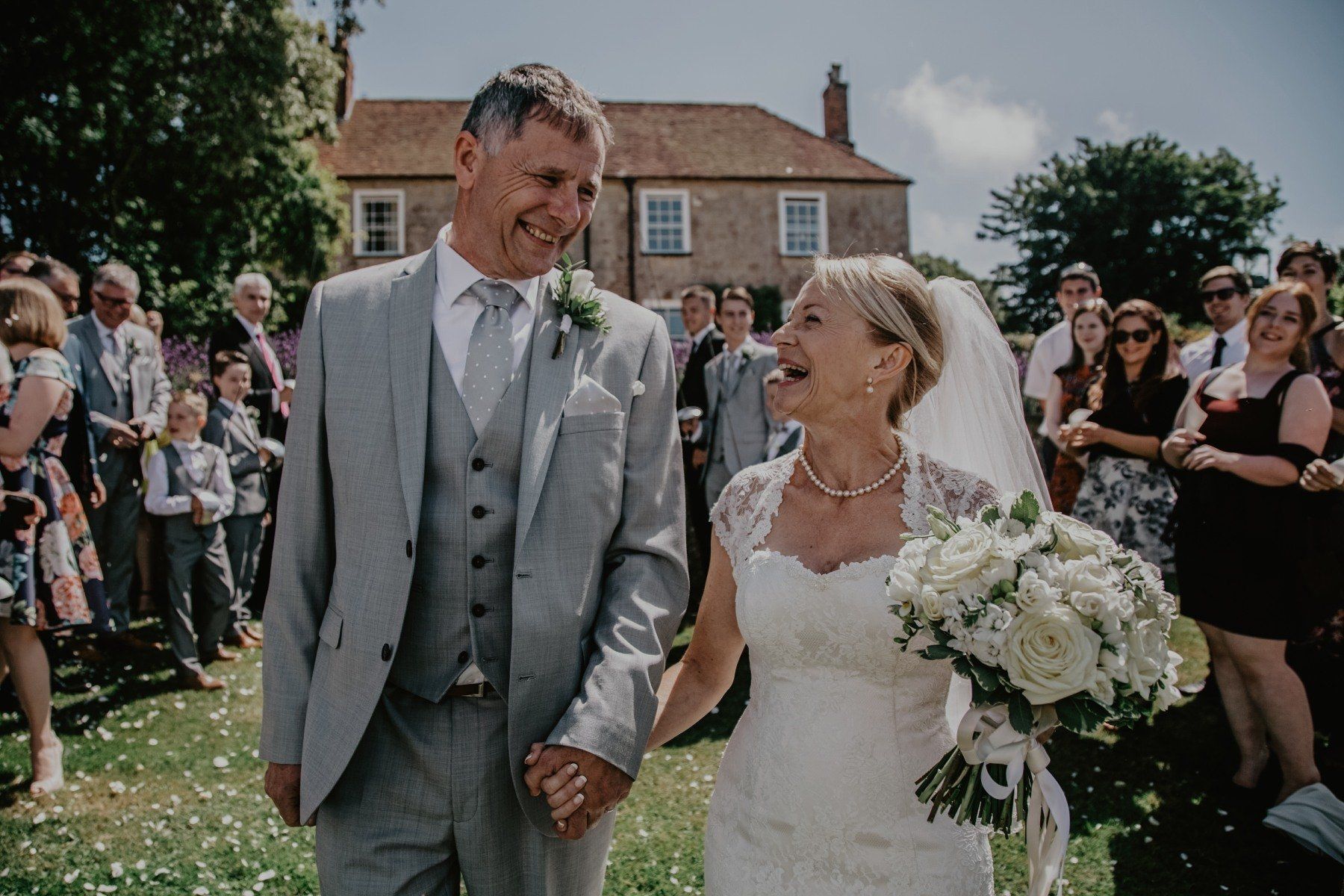 Newlyweds walk hand-in-hand, smiling, surrounded by guests. The man wears a grey suit, the woman a white dress.