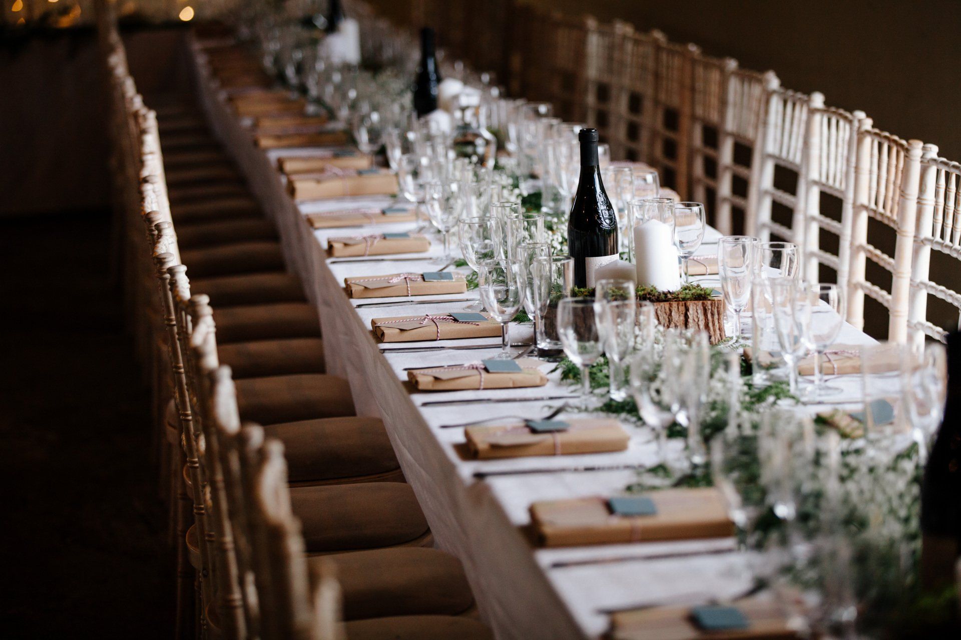 Long, decorated dining table set for a wedding; chairs on both sides, centerpieces, wine bottles.