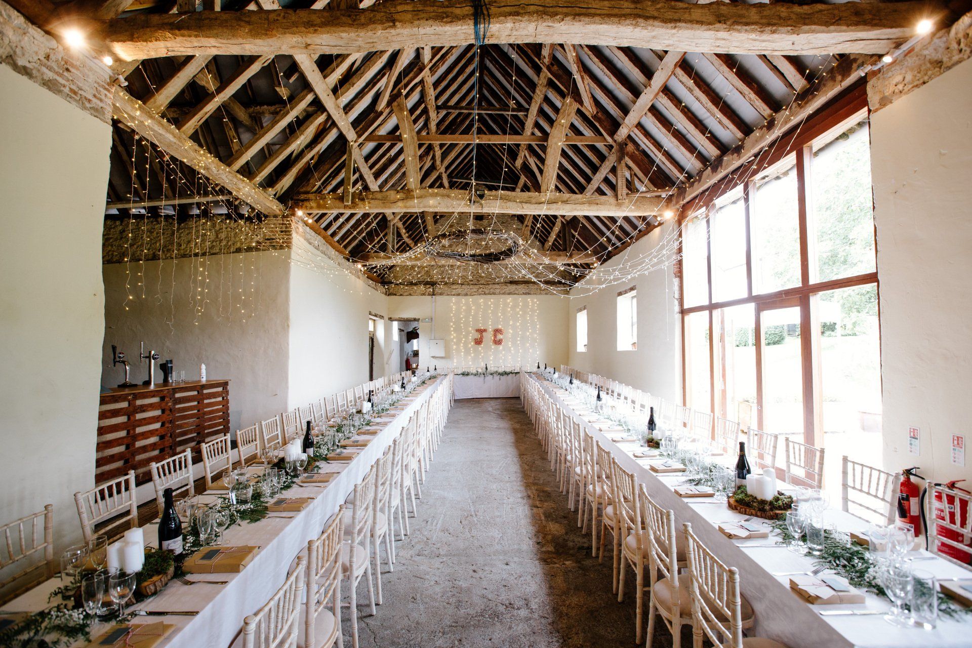Long, formal dining tables in a rustic barn with string lights, set for a wedding.