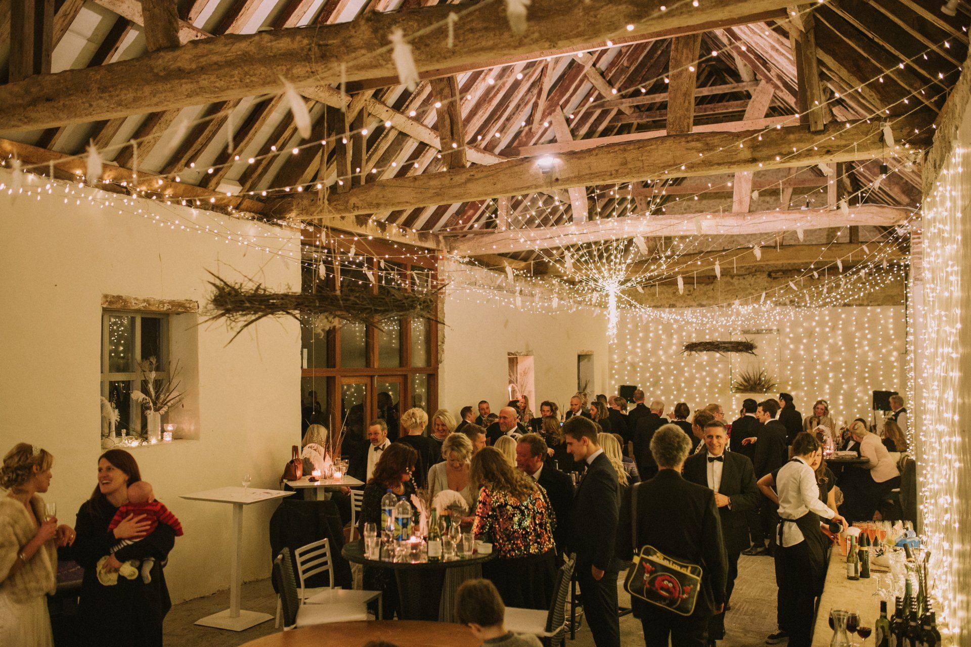 People mingle inside a warmly lit barn, adorned with string lights and rustic beams.