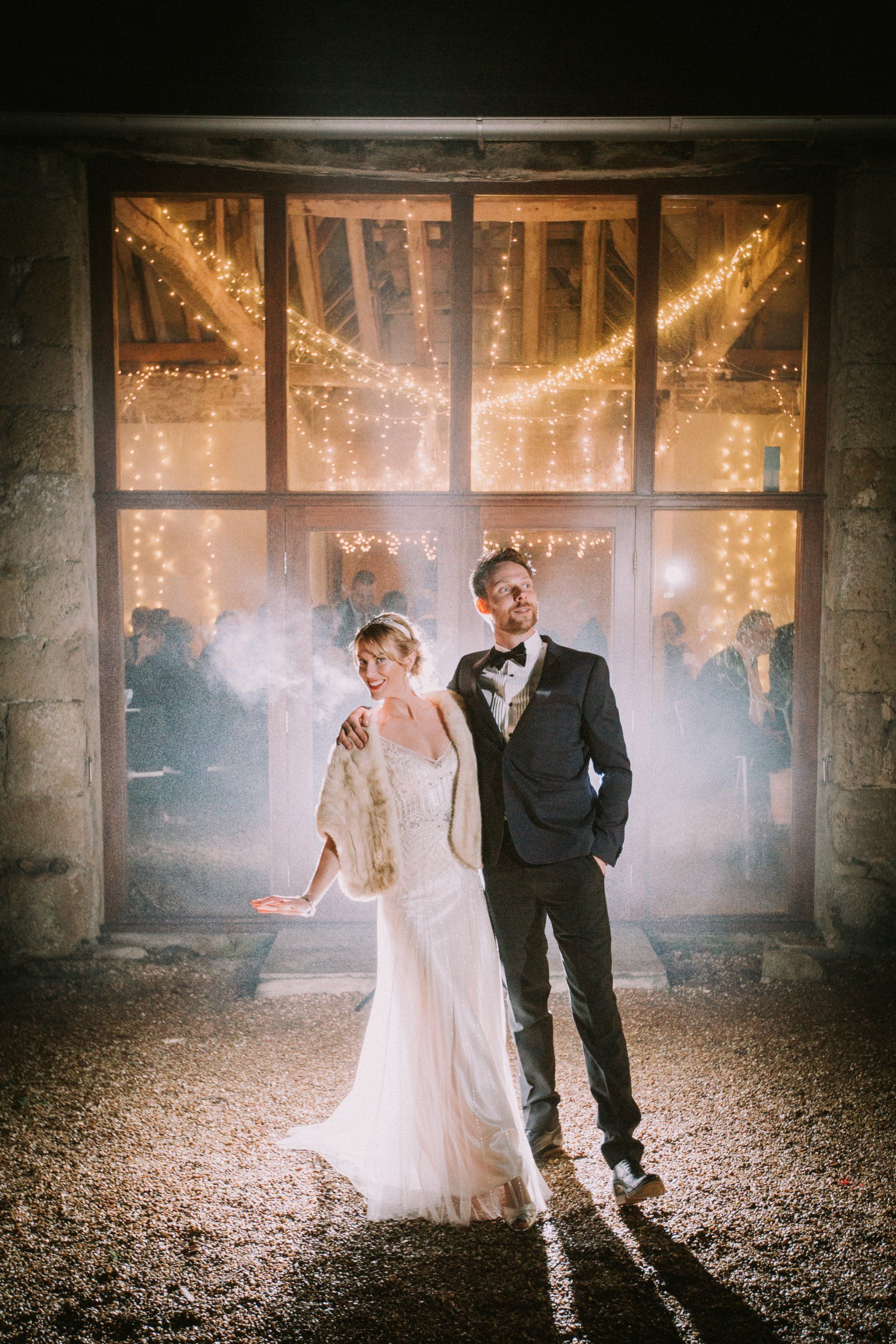 Newlyweds posing outside a lit building. The bride wears a gown and shawl, the groom a suit. Smoke and lights present.