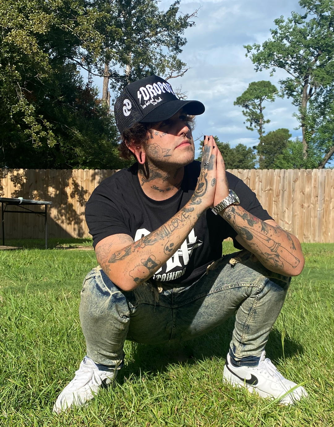 A man with tattoos is squatting in the grass with his hands folded in prayer.