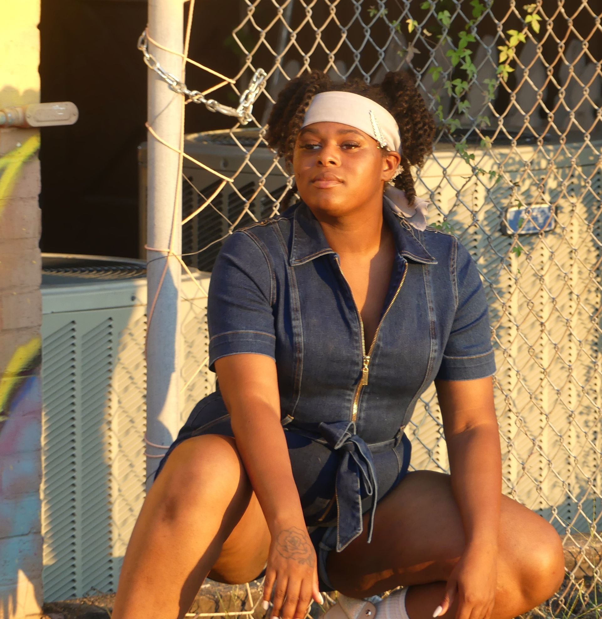 A woman in a denim dress is squatting down inf front of a hurricane fence.