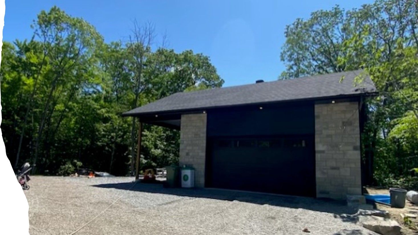 A garage with a black door is surrounded by trees and gravel.
