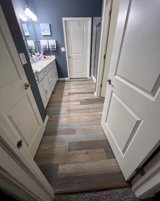 Narrow hallway with wood-look flooring leading to a blue bathroom with white cabinets and doors.