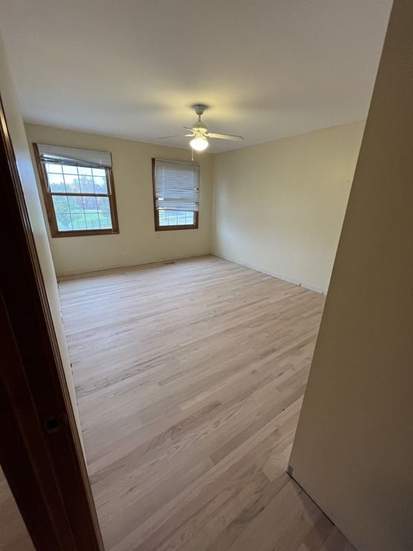 Empty beige bedroom with two windows, ceiling fan, and light wood flooring.