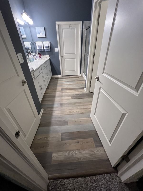 Narrow bathroom hallway with open white doors, gray wood-look floor, and vanity visible at left.