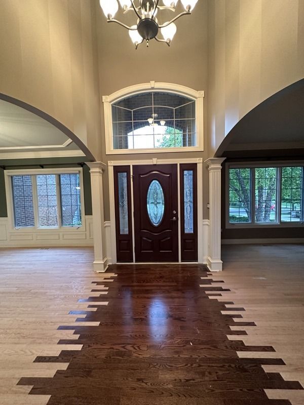 Empty foyer with dark double front doors, arched windows, and hardwood floor inlaid with light wood accents