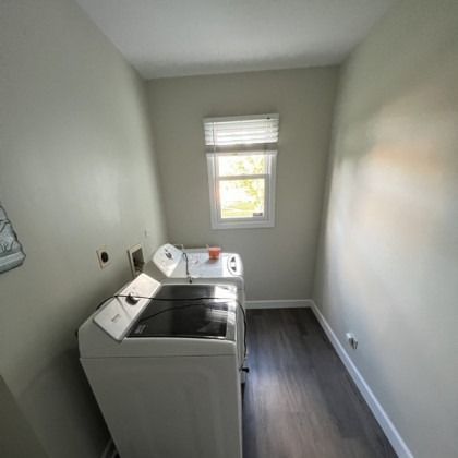 Narrow laundry room with washer and dryer, gray walls, dark wood floor, and a small window.