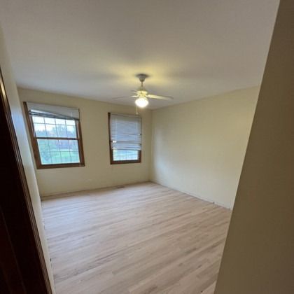 Empty beige bedroom with light wood floor, ceiling fan, and two windows.