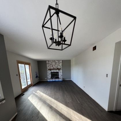 Empty modern living room with dark wood floors, geometric black chandelier, and stone fireplace at the far end