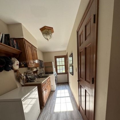 Narrow kitchen hallway with wooden cabinets, white counters, and sunlight on the tile floor