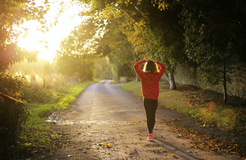 A woman is walking down a road with the sun shining through the trees.