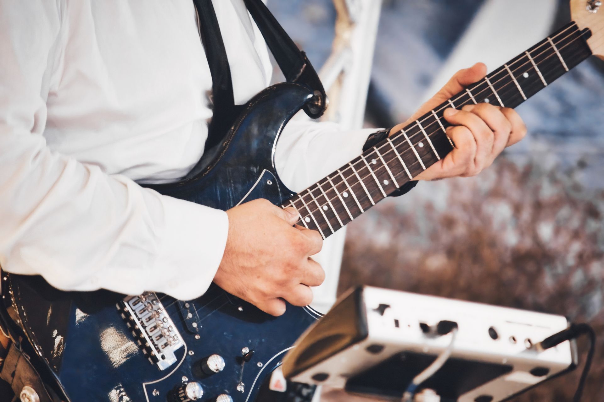 Guitarist playing a black electric guitar, close-up on hands and instrument. White shirt, stage lighting, amplifier.