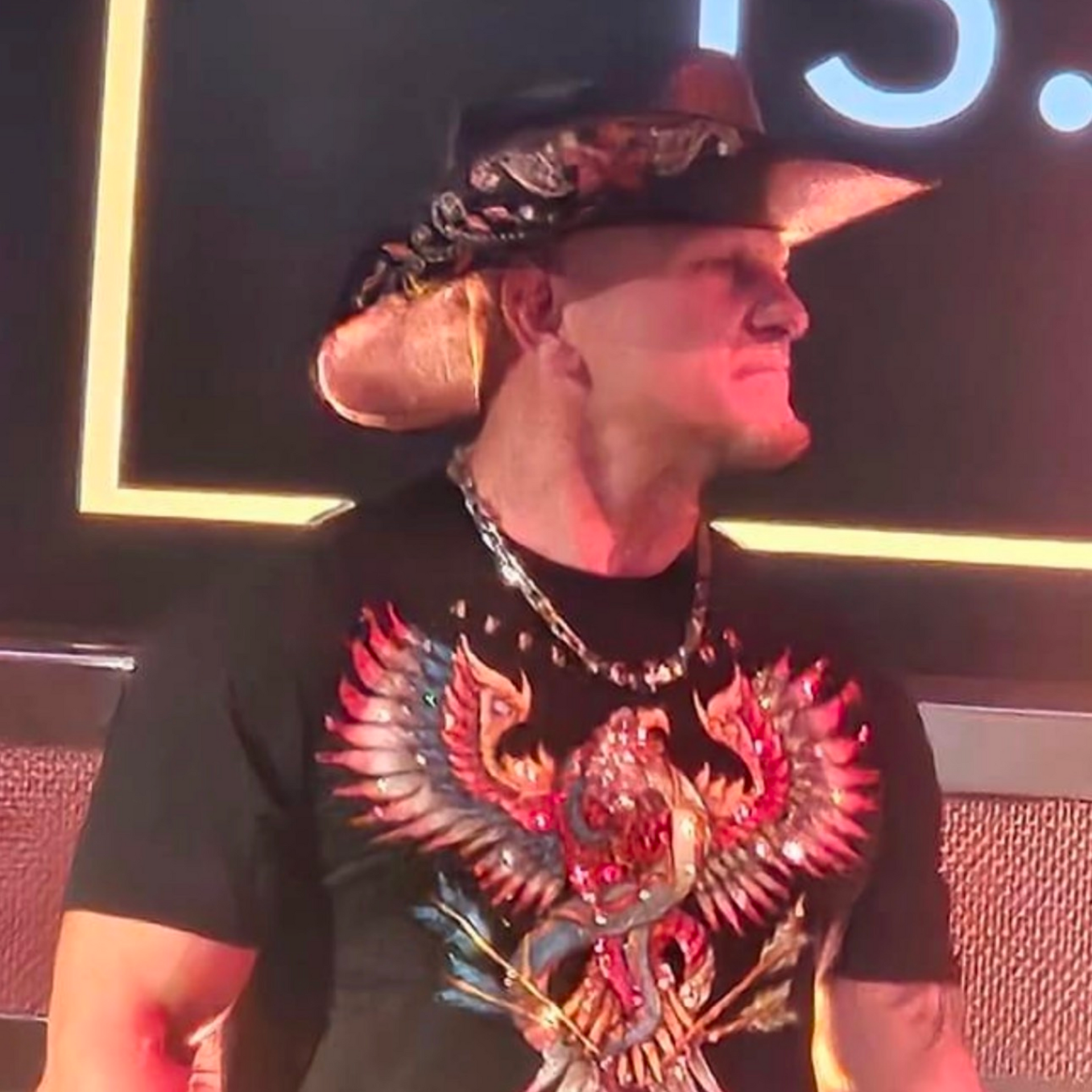 Man in cowboy hat and embellished shirt, with a chain necklace, in a dark setting, looking right.