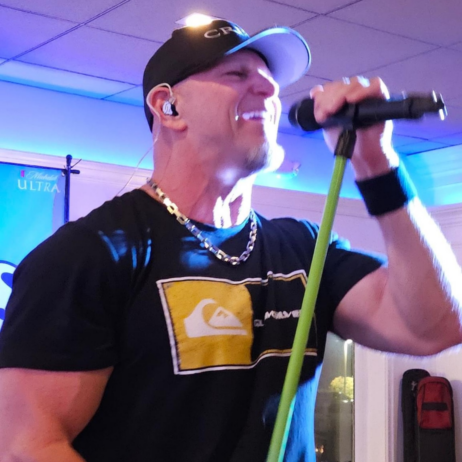 Man singing into a microphone, wearing a black cap and shirt, lit stage background.