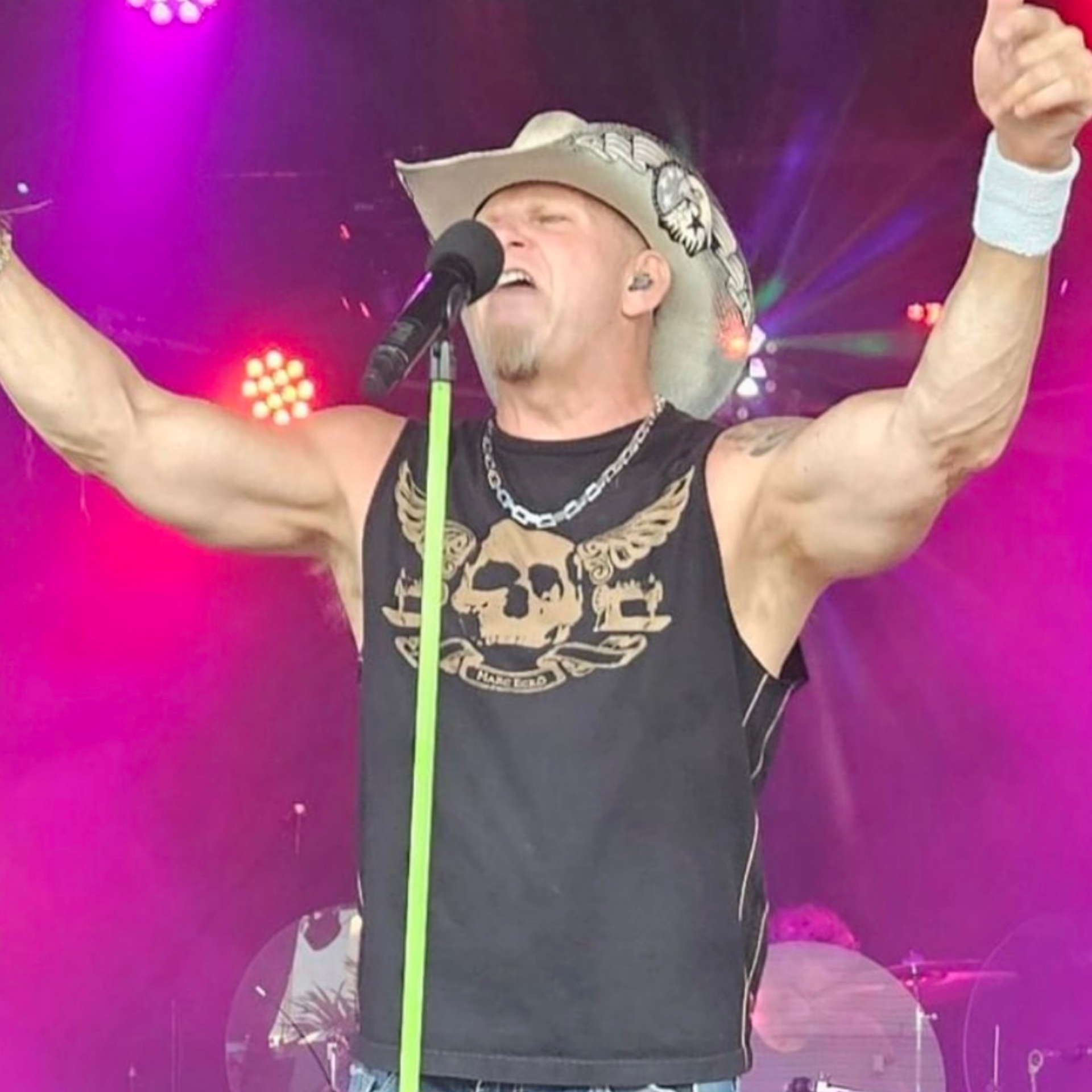 Man in cowboy hat sings into microphone, arms raised, on stage with pink and purple lights.