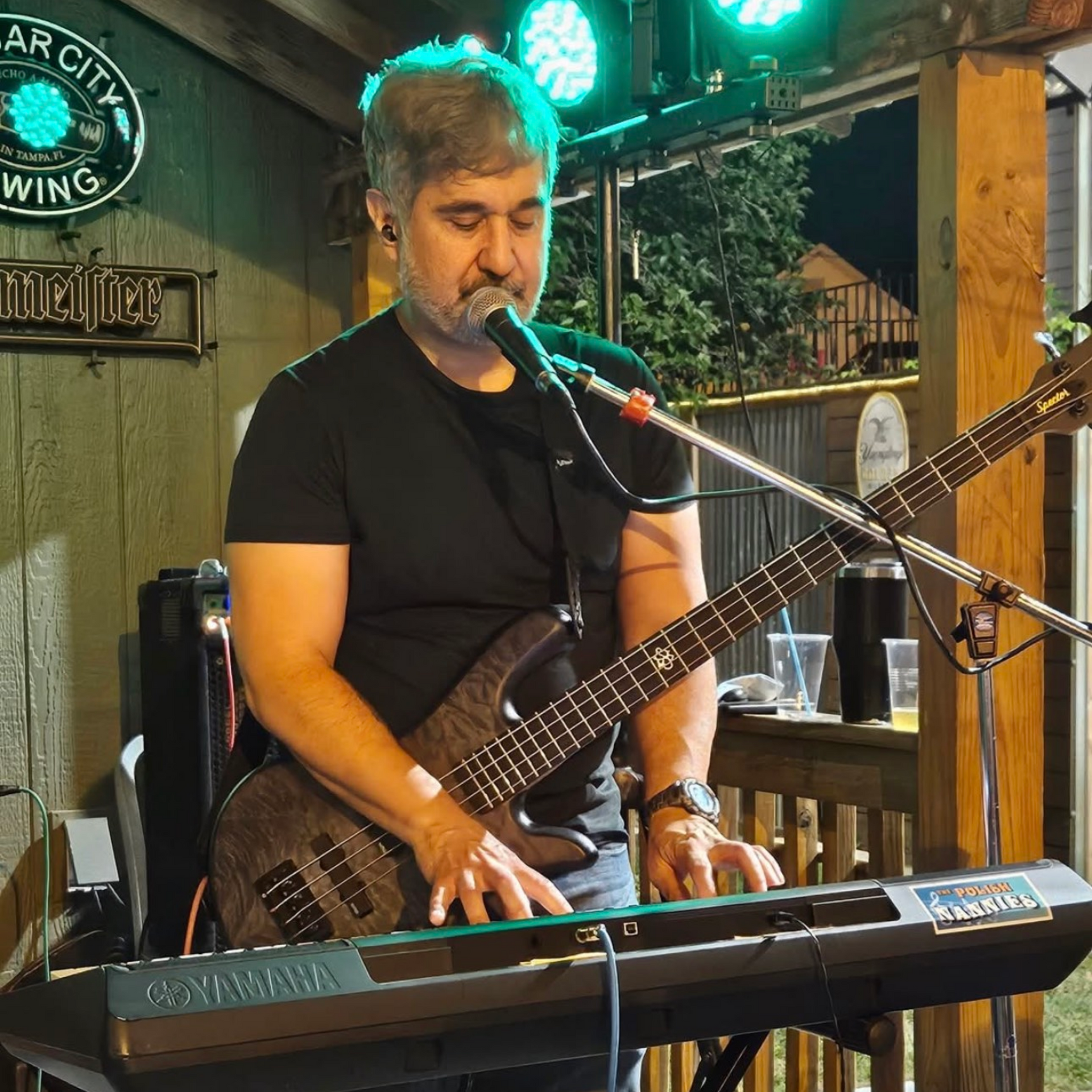Man playing a bass guitar and keyboard on an outdoor stage, under green and blue lights.