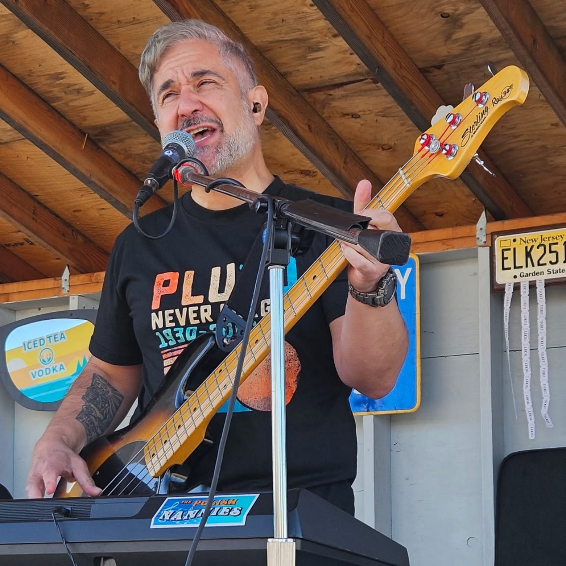 Man singing and playing bass guitar and keyboard on stage, wearing a black shirt, outdoors.