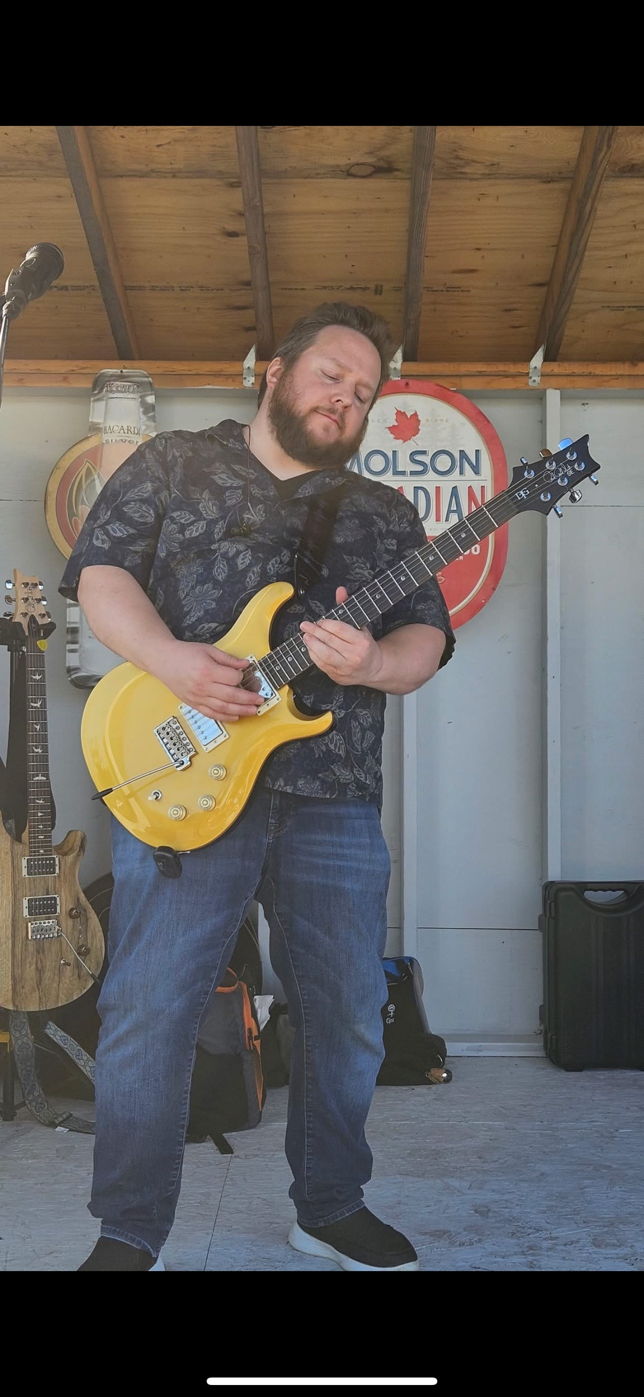 Man playing a yellow electric guitar outdoors. Behind him is a sign that reads