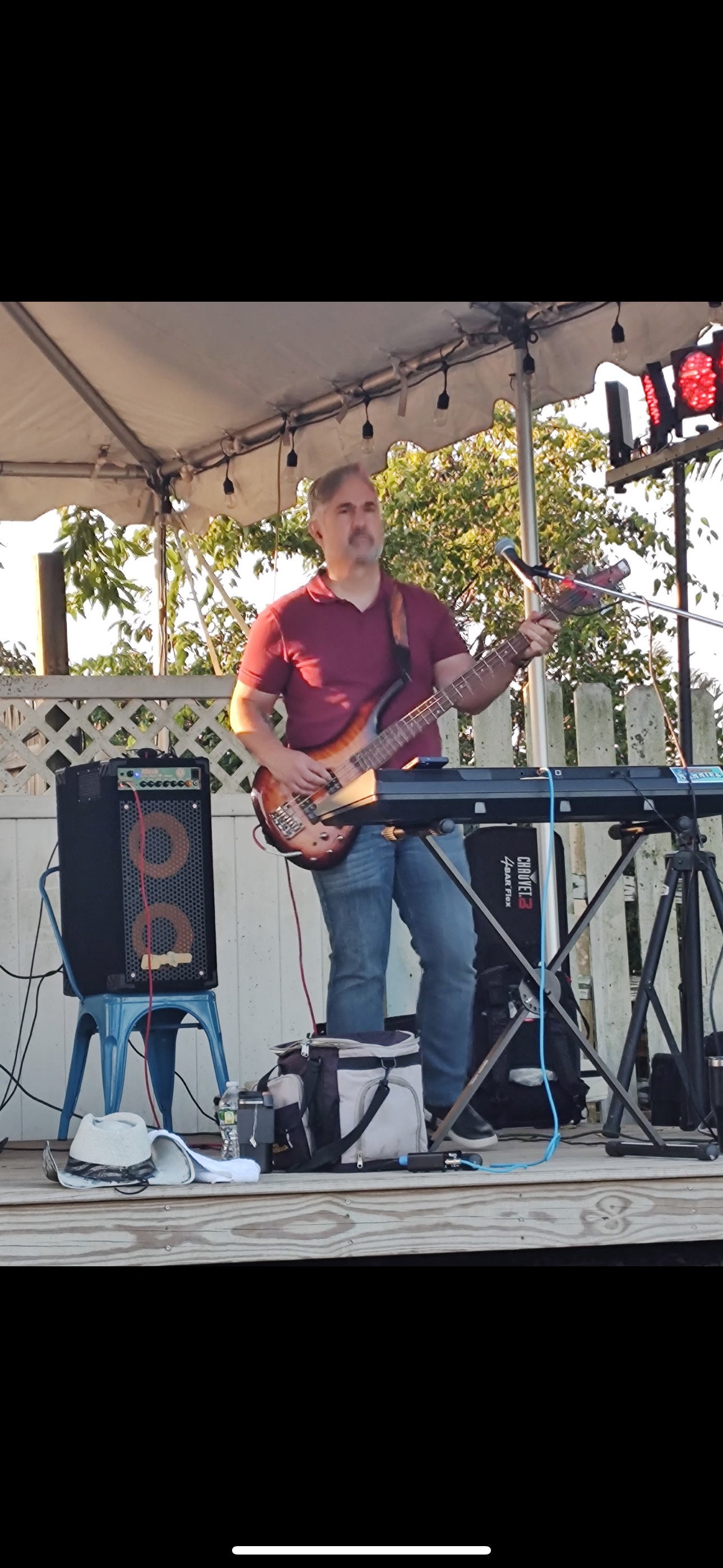 Man playing bass guitar on stage, with keyboard, amp, and outdoor setting.