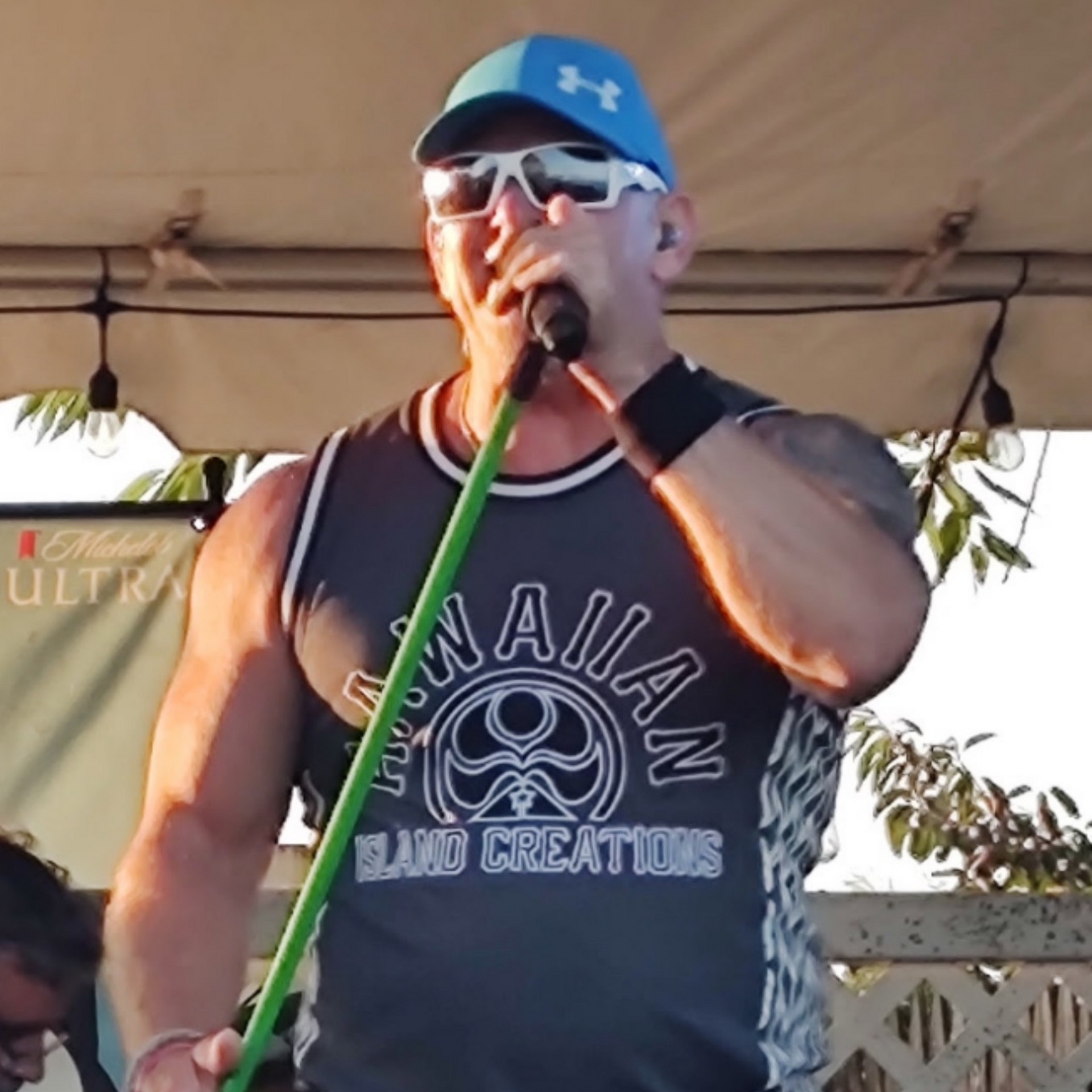 Man in blue cap and sunglasses singing into a microphone on stage, wearing a