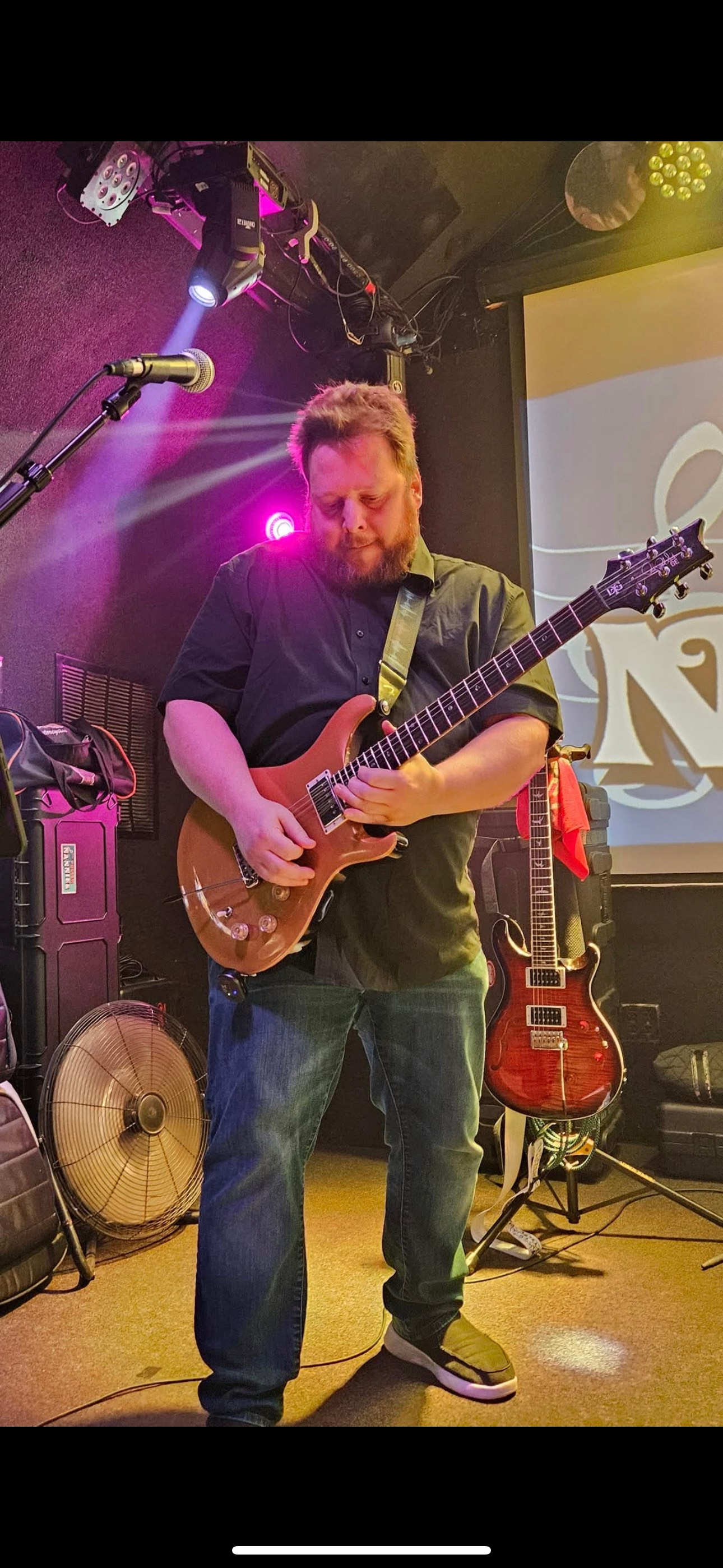 A musician playing a brown electric guitar on stage, illuminated by pink and purple stage lights.