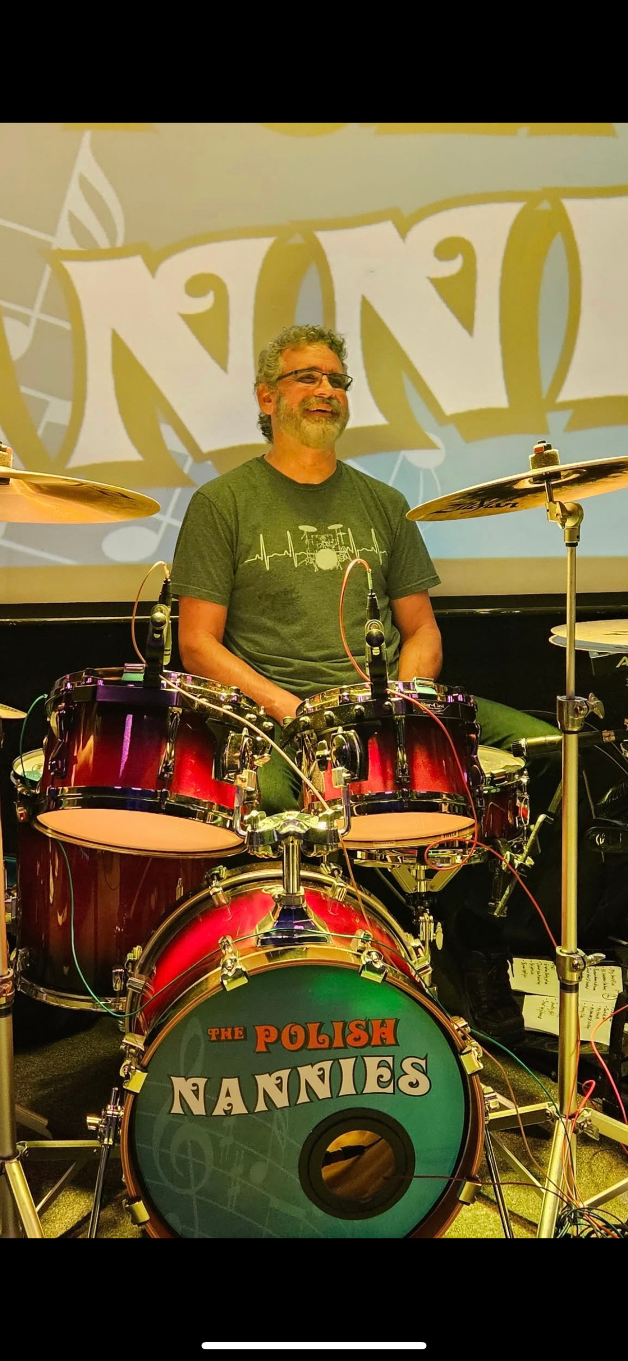 A drummer playing a drum kit onstage. The bass drum says,