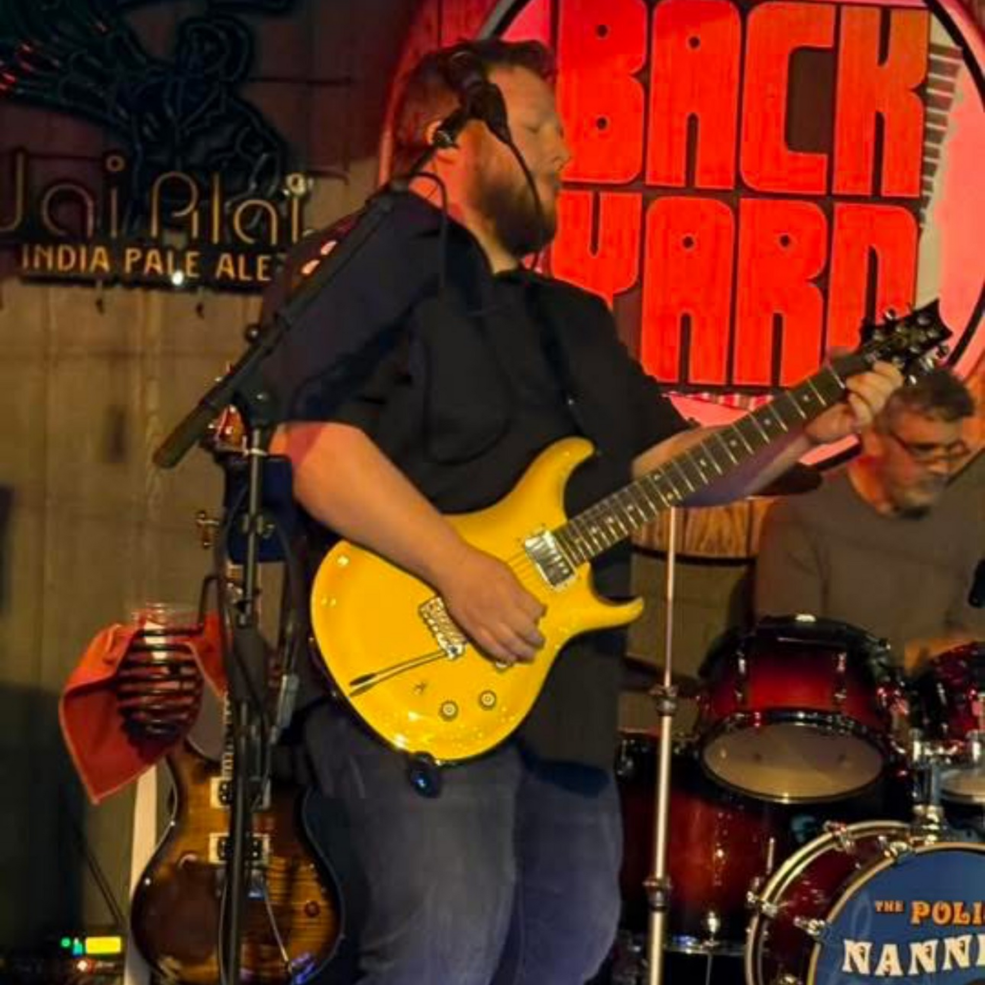 Guitarist on stage, playing a yellow electric guitar; drums and bassist visible;