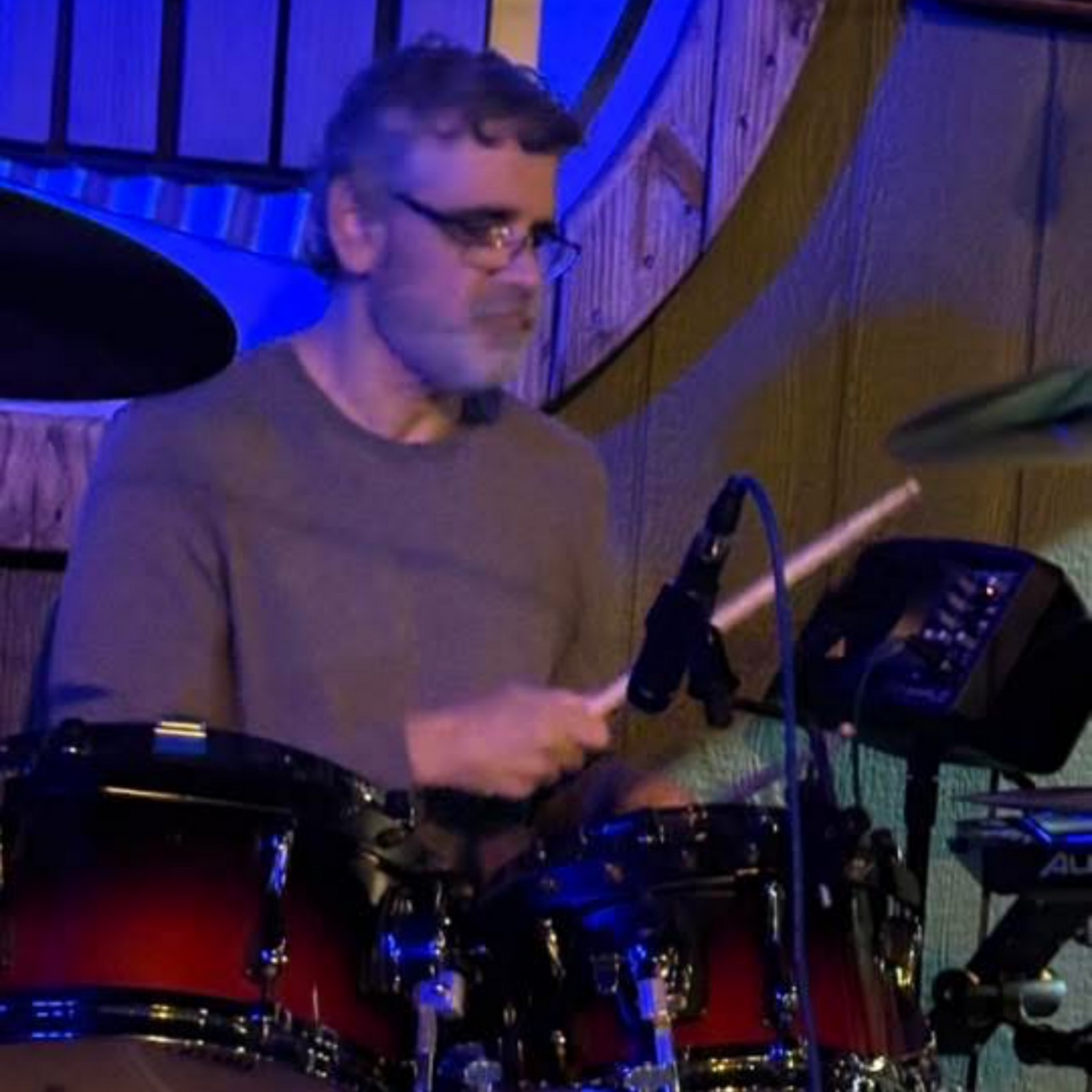 Man playing drums in dimly lit setting, holding drumsticks. The drums are red and black.