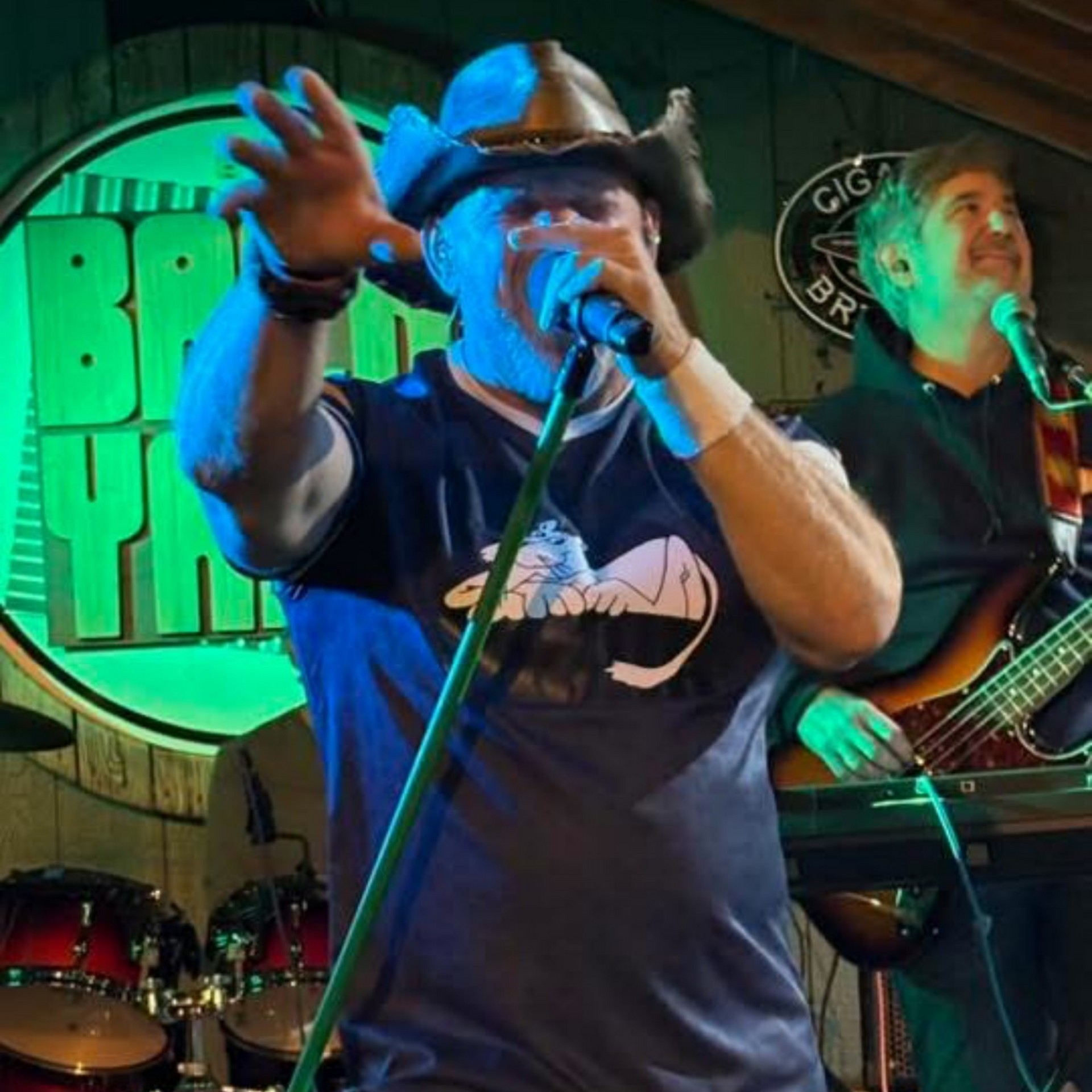 Man in cowboy hat singing into a microphone on stage, another person playing bass guitar.