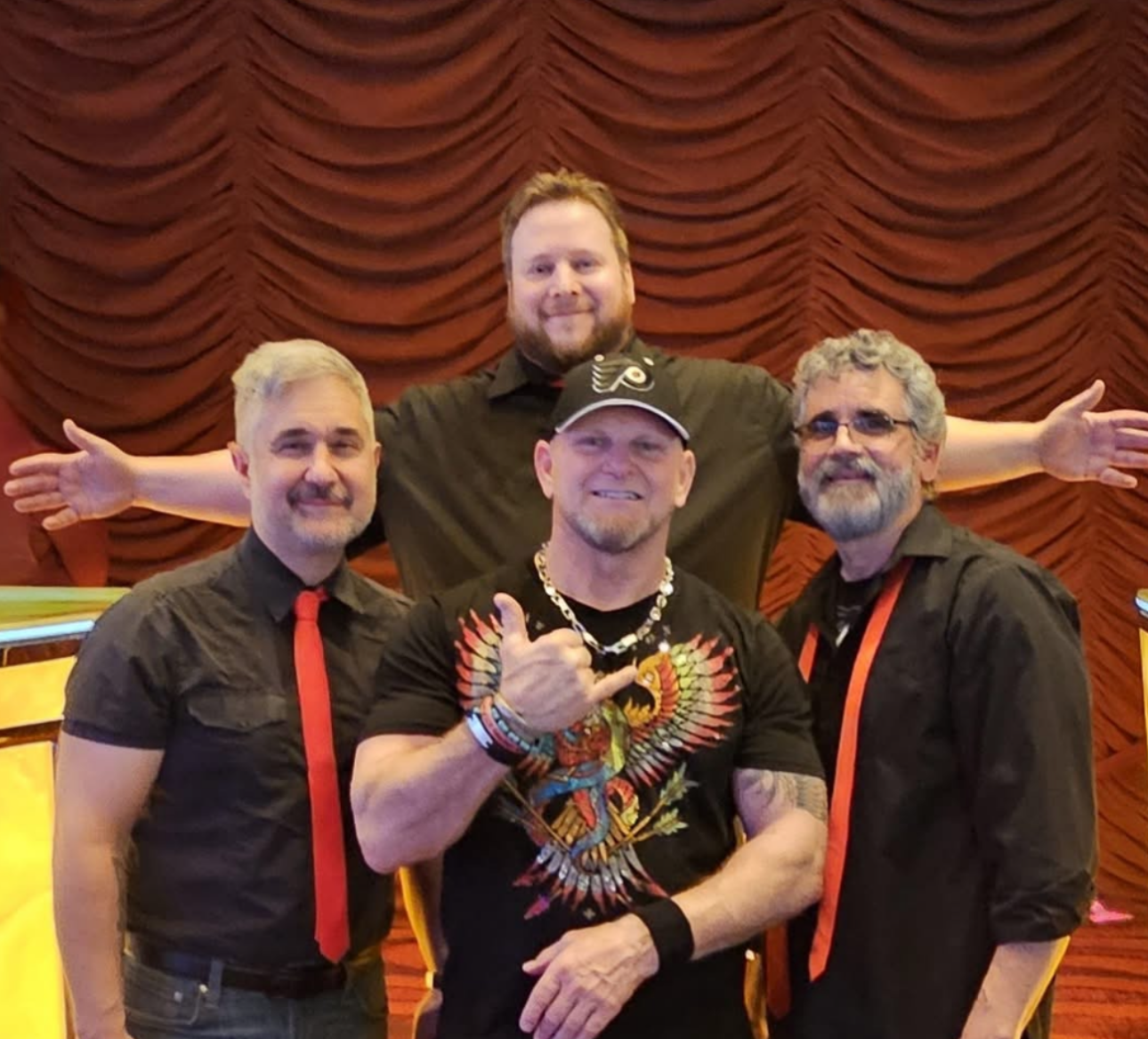 Four men pose for photo. One gives thumbs up. All are smiling. Red background.