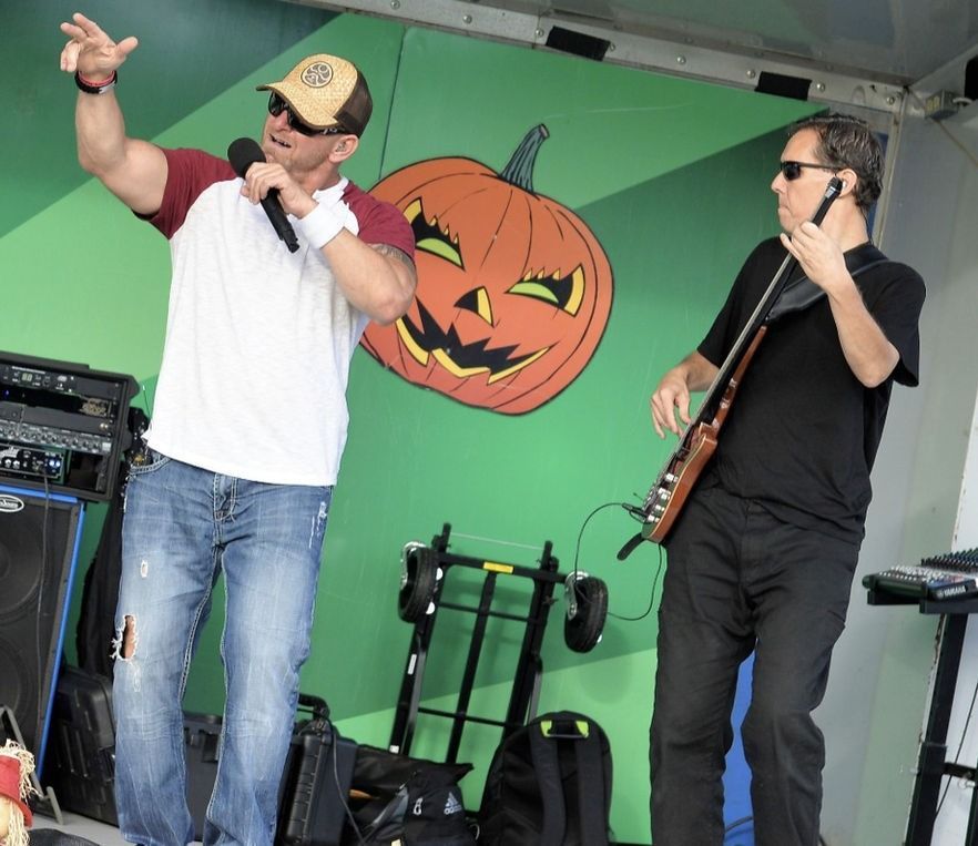 Two men perform on stage with a pumpkin backdrop. One sings, the other plays a bass guitar.