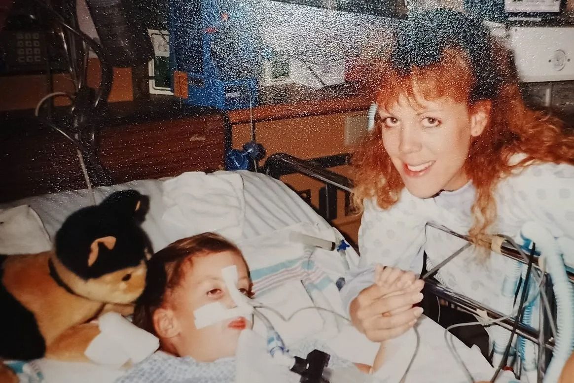 Person smiling beside a child in a hospital bed, with a stuffed animal and medical tubing visible.