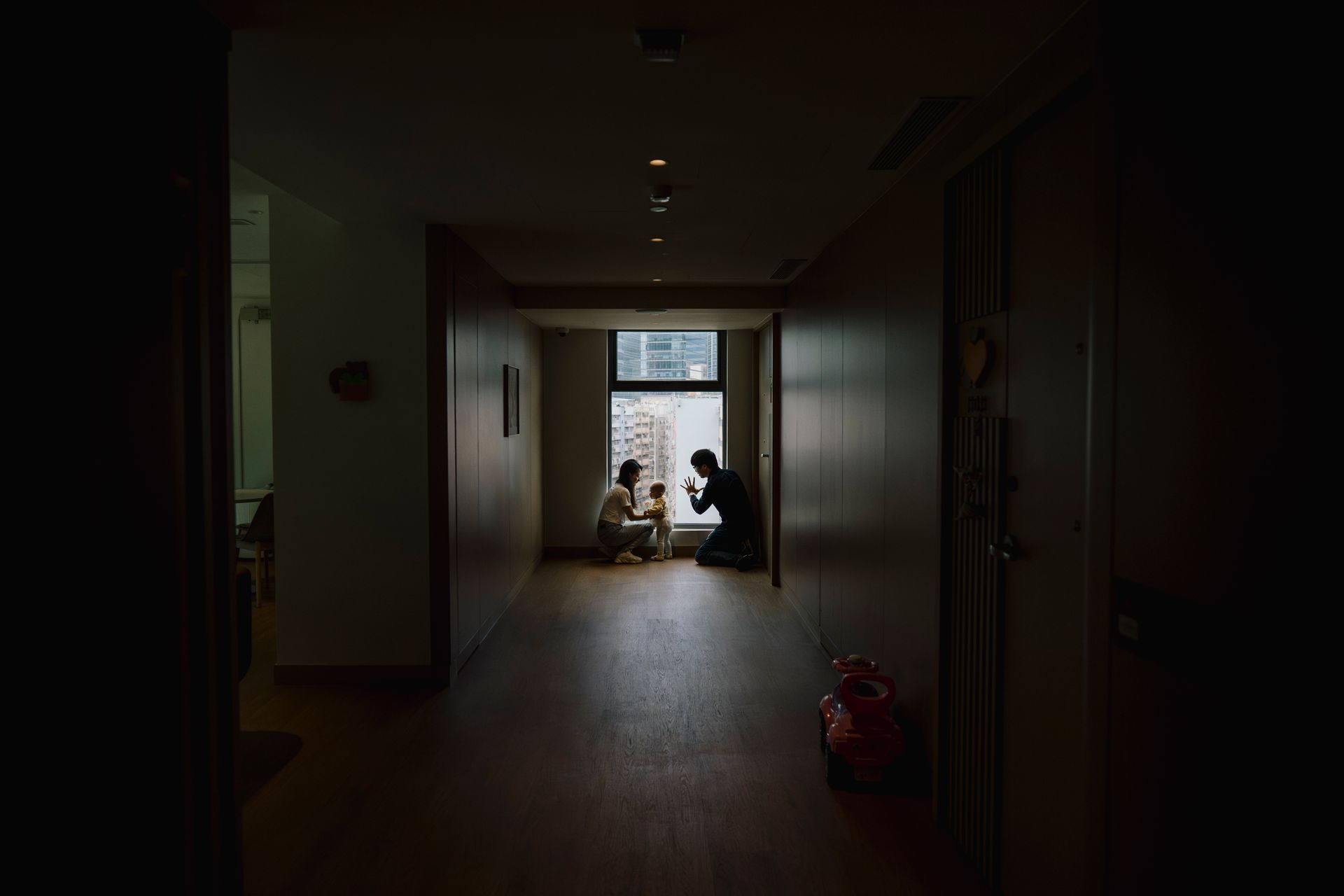 Dark hallway with two people seated at the far end near a bright window, one crouching beside them.