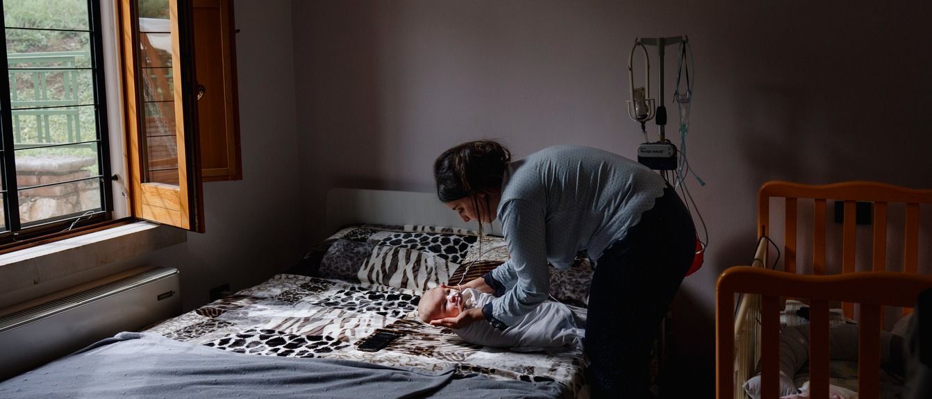 Person bending over a bed, making it up in a dim bedroom near an open window.