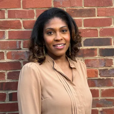 Smiling person in a beige blouse standing against a red brick wall