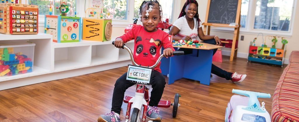 Child riding a tricycle in a colorful classroom, with toys and another child in the background