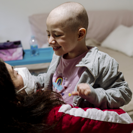 Smiling child in a gray hoodie sitting on a bed and playing with an adult in a red striped top