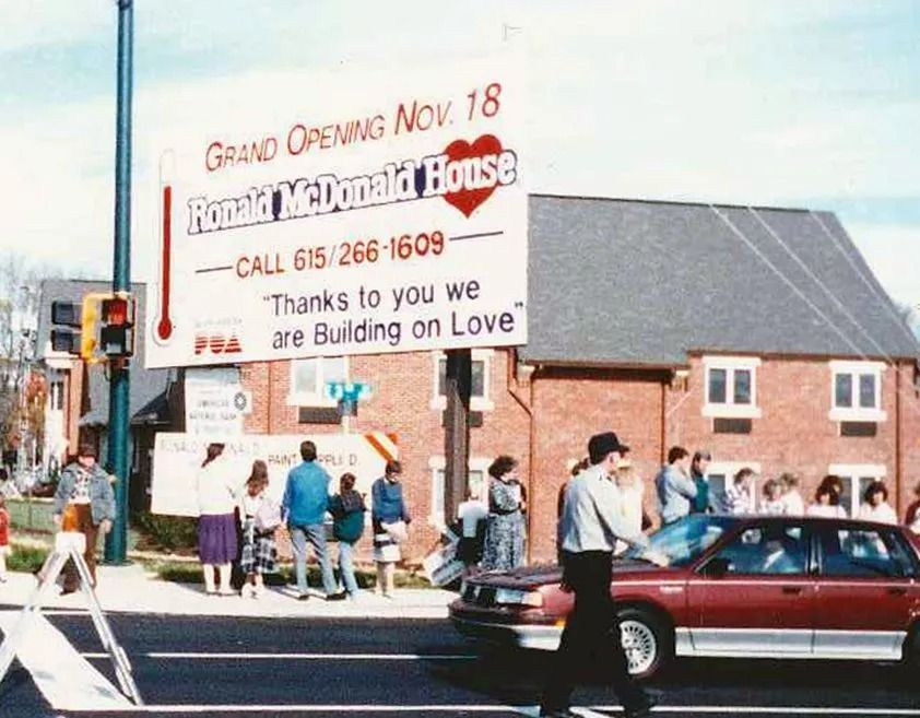 Grand opening sign outside a red brick building, with people gathered on the sidewalk and cars passing by.